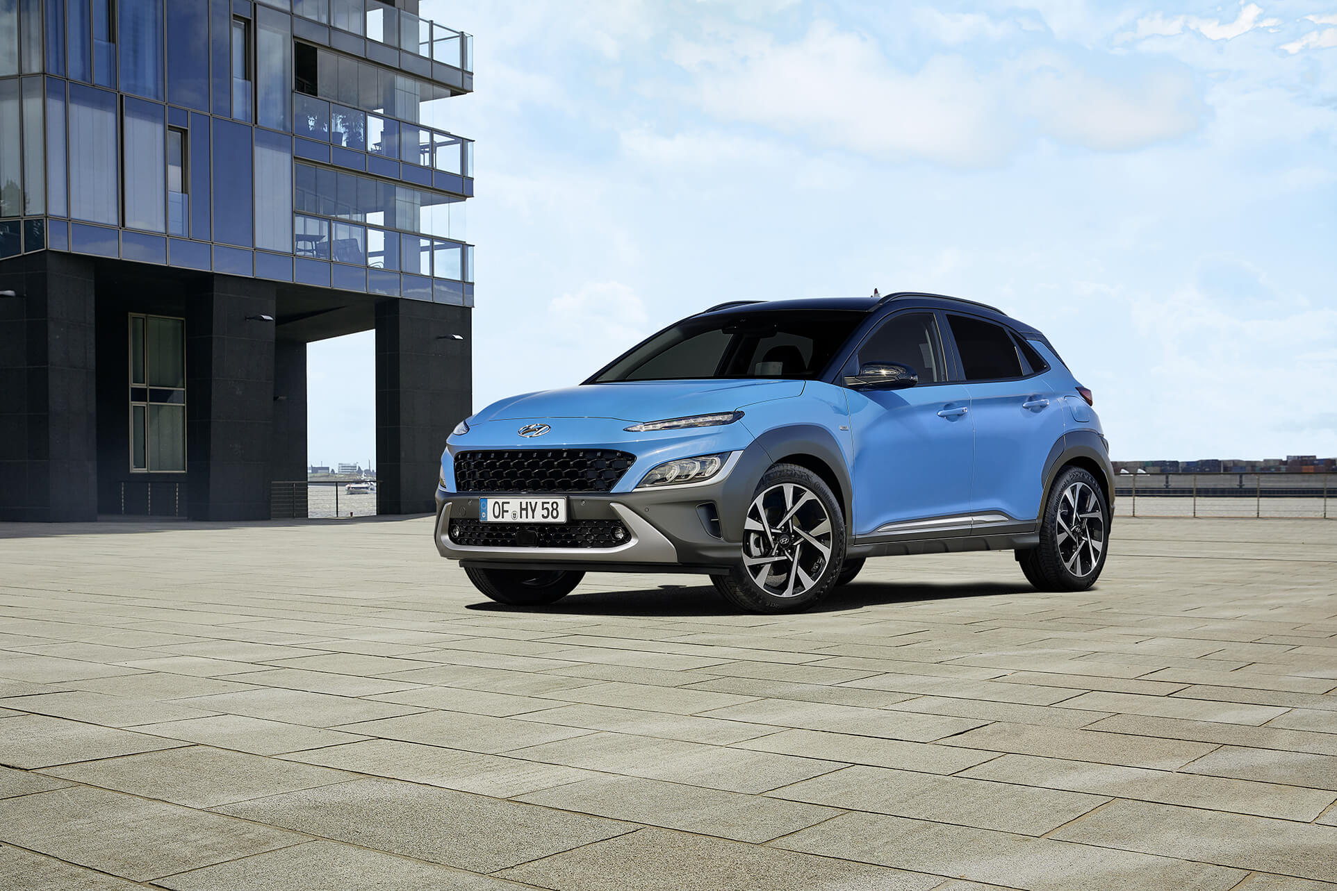 Nový Hyundai Kona v barvě Surfy Blue, zaparkovaný na otevřeném náměstí, před moderní prosklenou budovou.